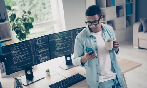 A business owner looking at his phone while standing in front of his computer desk.