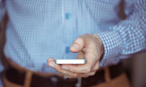 A close-up of a person in a blue checkered shirt and brown belt holding a smartphone, focusing on their hand and the device.