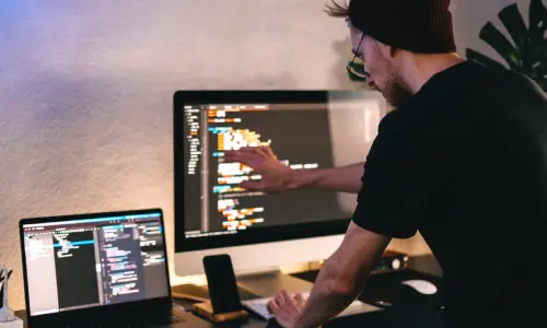 A rear-view shot of a computer programmer in a black shirt working on multiple electronic devices at the same time.