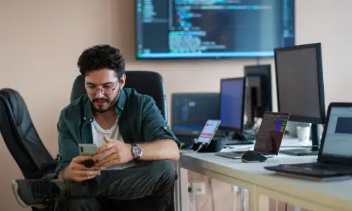 A computer programmer on his phone and seated next to a desk where he is working on multiple electronic devices.
